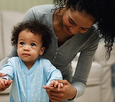 woman bent over holding hands of toddler learning to walk