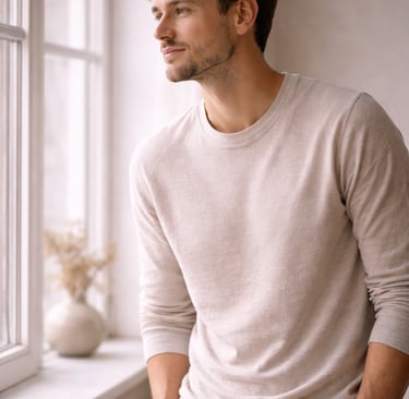A handsome man in a beige crew neck sweater looking out a bright window.