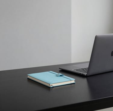 A minimalist studio desk in a South American / Brazilian office, featuring a sleek black surface, a silver laptop, and a small baby blue decorative notebook. The lighting is soft and natural, emphasizing clean dark gray and light gray lines in a professional setting.