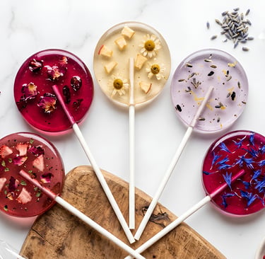 Handmade gourmet lollipops with edible flowers and herbs on a marble background.
