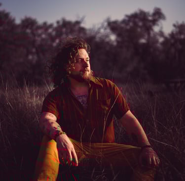 Artistic portrait of a man with long wind-swept hair sits in long grass at sunset