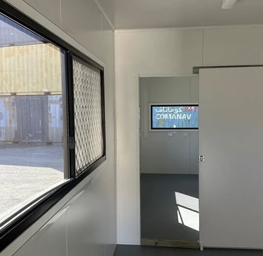 Interior of a white portable site office with a sliding door and windows overlooking a shipping container yard.
