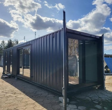 Modern dark grey shipping container home with large glass sliding doors under a bright blue sky.