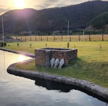 Sunset over a tranquil mountain pond with a stone barbecue pit on a lush green lawn.