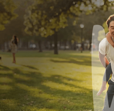 A happy father gives his young daughter a piggyback ride in a sunny, green park.