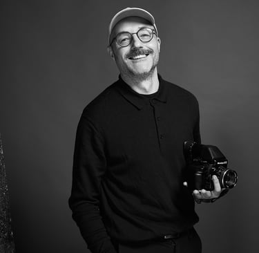 A smiling photographer in a cap holding a professional camera against a grey background.