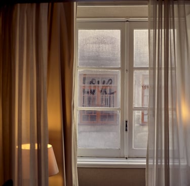 The word love written on a foggy glass window pane framed by white curtains and warm lamp light.