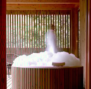 A rustic round cedar hot tub filled with white bubbles on a wooden patio with slatted privacy walls.