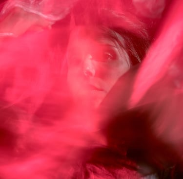 Dramatic artistic portrait of a woman peering through translucent layers of vibrant red fabric.