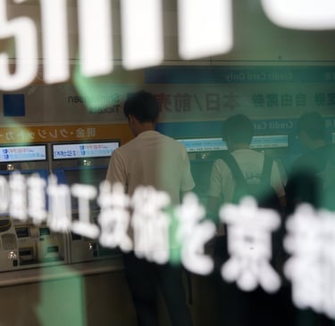 Commuters buying tickets at automated train station kiosks in Japan through a glass window.