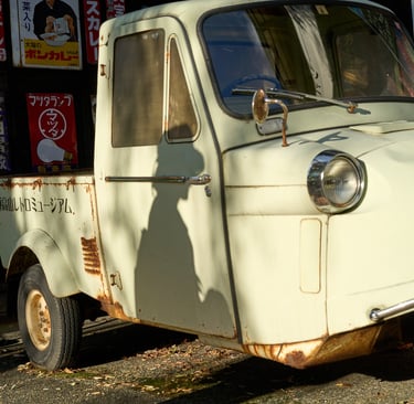 Vintage white Daihatsu Midget three-wheeled truck parked in front of retro Japanese metal signs.