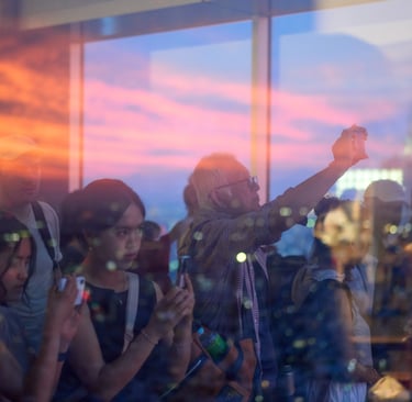 People taking photos of a colorful sunset from a glass observation deck overlooking a city skyline.