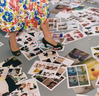 A person in a floral dress and green heels stands among many photographic prints scattered on a studio floor.