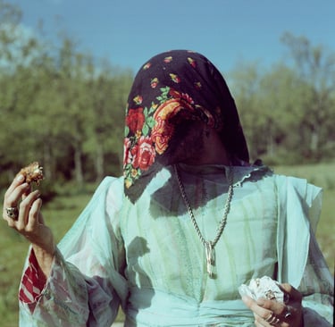 Person in vintage floral headscarf and light blue gown holding food in an outdoor setting.