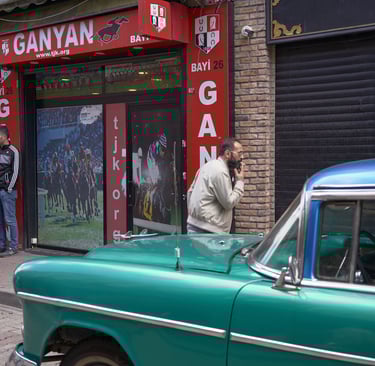 Vintage green classic car parked in front of a red Ganyan horse racing betting shop on a city street.