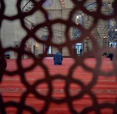A man praying on a red carpet inside a mosque, viewed through an ornamental geometric screen.