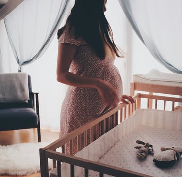 A pregnant woman stands in a sunny nursery, holding her baby bump next to a wooden crib.