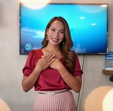 Smiling woman in red silk blouse and pink pleated skirt posing with hands over heart.