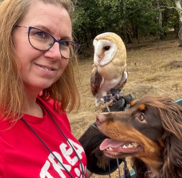 Dog trainer smiling with her spaniel and a barn owl perched on her fist outdoors.