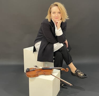 Violinist Žydrė sitting in a studio, leaning forward with hand under chin, violin resting beside her