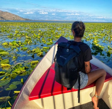 A traveler wearing a black waterproof backpack sits on a boat in a lake covered in water lilies.