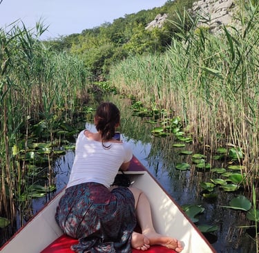 A woman sits in a boat on a narrow river path surrounded by tall reeds and lily pads.