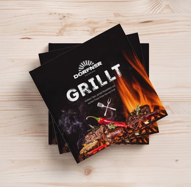 A stack of Dorfner Catering Grillt cookbooks featuring a flaming barbecue cover on a light wood background.