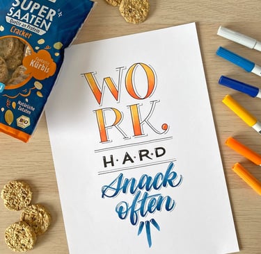 Hand-lettered motivational quote on a desk with colorful markers and healthy pumpkin seed crackers.