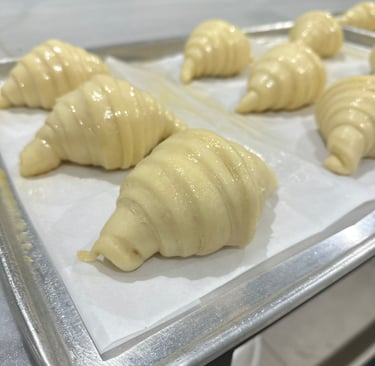 Freshly rolled raw croissant dough resting on a baking sheet with parchment paper in a professional bakery.