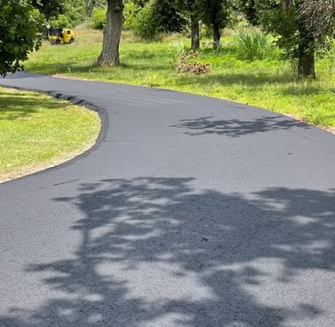 Freshly paved black asphalt driveway winding through a green lawn with residential landscaping and trees.