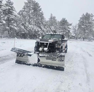 A black pickup truck with a SnowDogg v-plow clearing deep snow on a winter driveway.