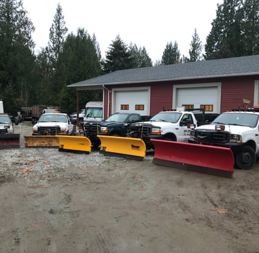 5 trucks with plows are parked in front of a 3 bay shop. The trucks are in a semi circle facing the camera