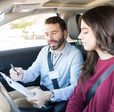 A professional driving instructor with a clipboard giving a student driver feedback inside a car.