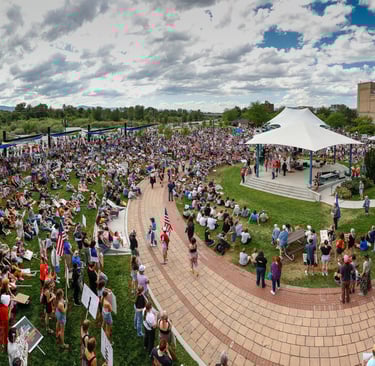 A large crowd gathers for a political rally and protest at Caras Park with a white tent stage.