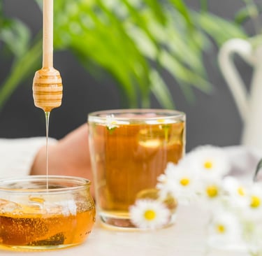 Golden organic honey dripping from a wooden dipper into a jar next to herbal chamomile tea.