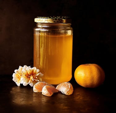 A glass jar of organic golden honey with fresh tangerine slices and flowers on a dark background.