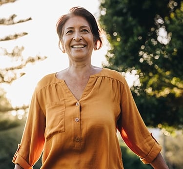 A smiling senior woman in an orange blouse enjoying a sunny day outdoors at a park.