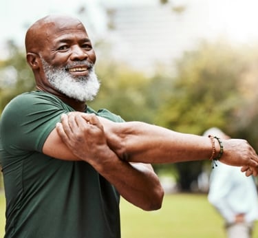 A smiling senior African American man with a white beard stretches his arm during an outdoor park workout.