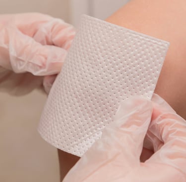 Gloved hands applying a white sterile adhesive wound dressing patch to a patient's leg for injury care.
