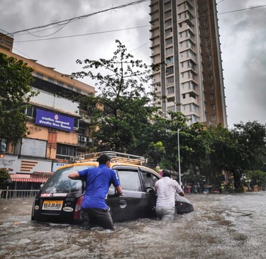 Men pushing a black taxi through deep flood water on a city street in Mumbai during heavy monsoon rain.