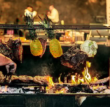 Chef asando chorizo argentino y carnes colgadas con piñas sobre un asador de leña al fuego abierto.