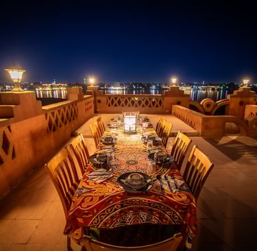 Elegant outdoor dining table set for dinner on a terrace overlooking the Nile River at night.