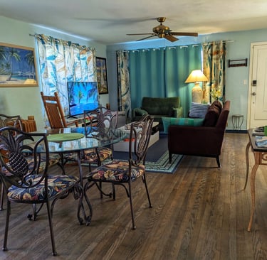 A tropical themed living and dining room with a glass table, dark hardwood floors, and teal curtains.