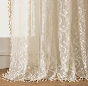 Elegant cream floral embroidered lace curtains with bohemian fringe tassels on a wooden floor.