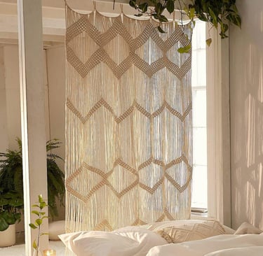 A decorative boho macrame wall hanging used as a stylish room divider in a bright, sunlit bedroom.