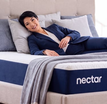 A smiling woman in a navy suit relaxes on a comfortable Nectar memory foam mattress with plush pillows.