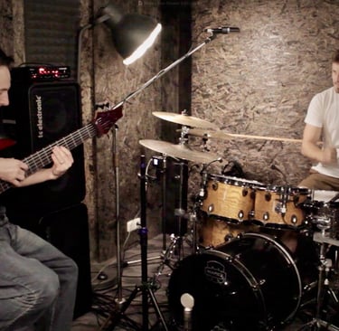 Jozef Bobula on red six-string bass with drummer in a rustic dimly lit rehearsal space.