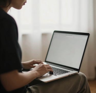 A person typing on a laptop computer while working from home near a bright window.