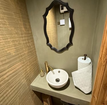 Rustic bathroom with white washbasin, gold tap and black mirror set against olive green microcement
