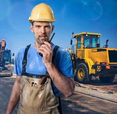 A construction worker in a yellow hard hat using a walkie-talkie at a roadwork site with heavy machinery.
